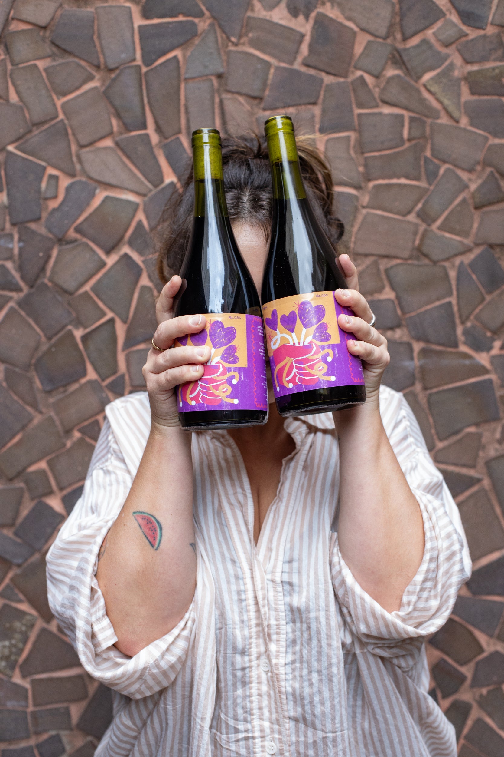 Foto de uma pessoa com uma camisa listrada, em frente a uma parede de pedras, segurando duas garrafas de vinho em frente ao rosto.